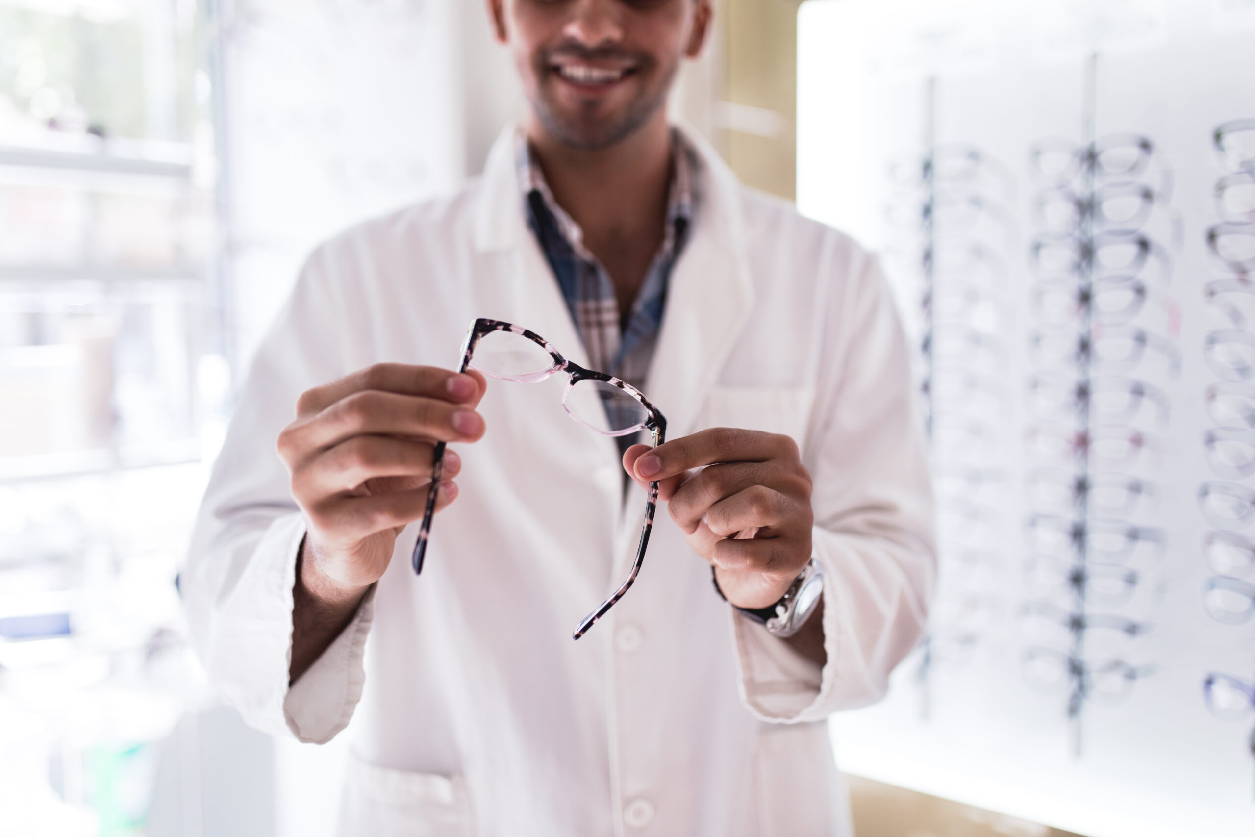 Handsome ophthalmologist holding eyeglasses frame in optical store. Selective focus on hand and eyeglasses.
